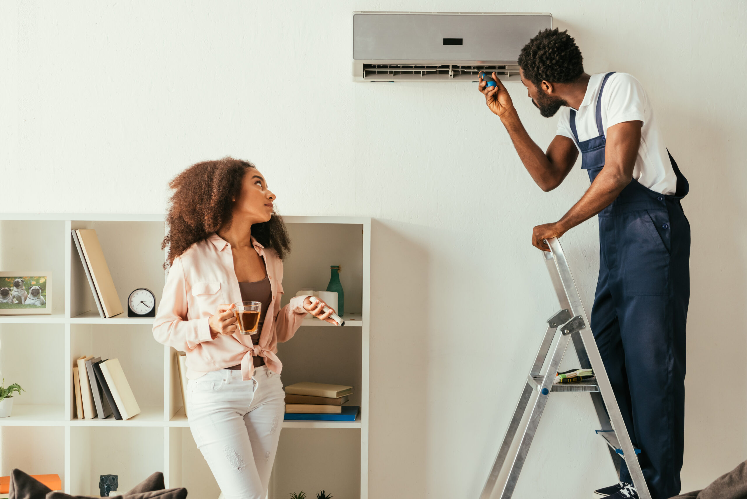 african american repairman fixing air conditioner near pretty african american woman holding remote